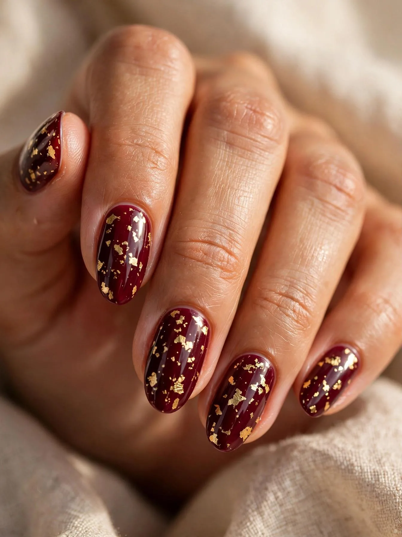 Medium almond burgundy nails with irregular hand-placed gold foil shards.