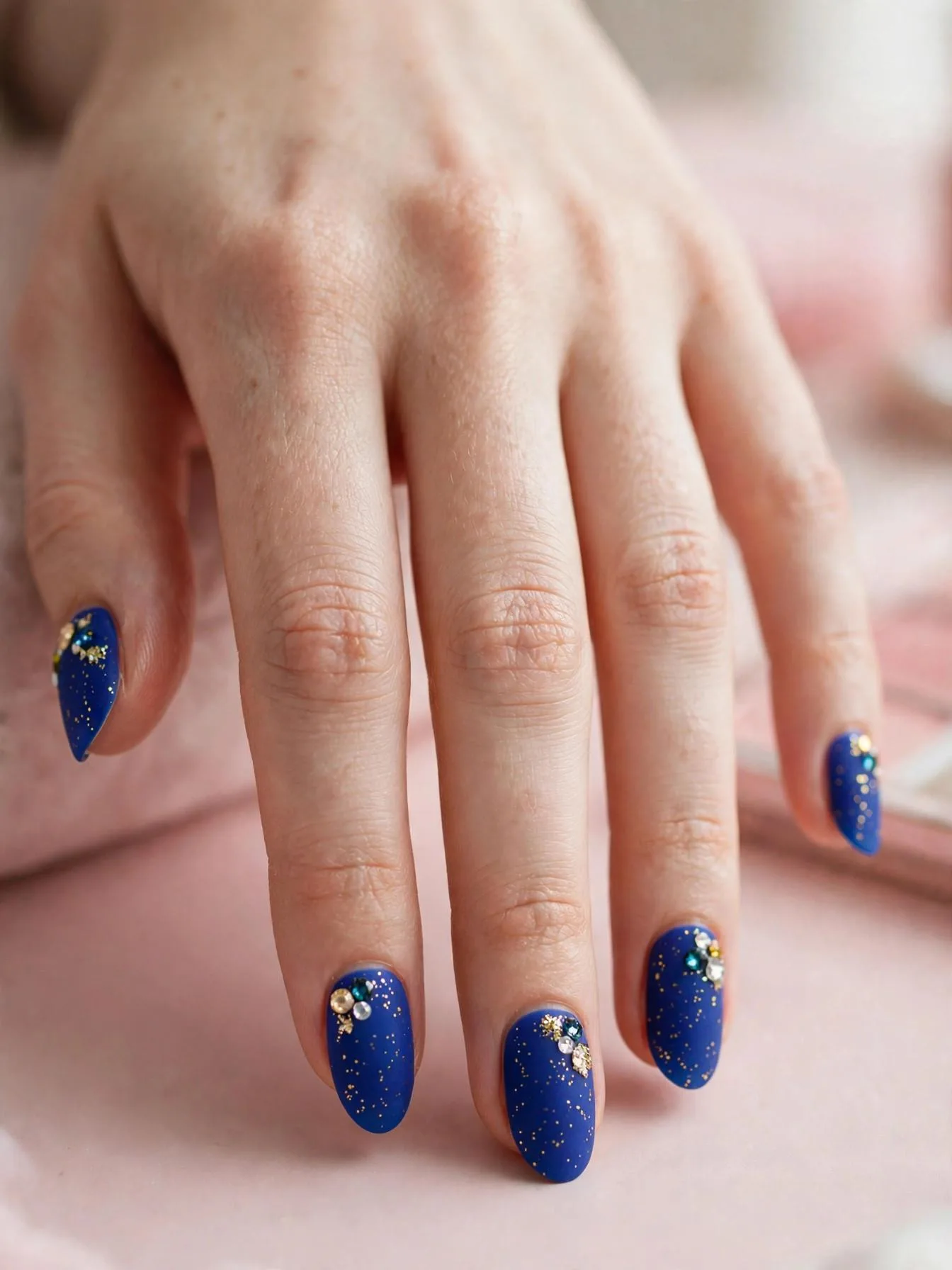 Medium almond nails in matte royal blue with gold glitter and small gemstones.