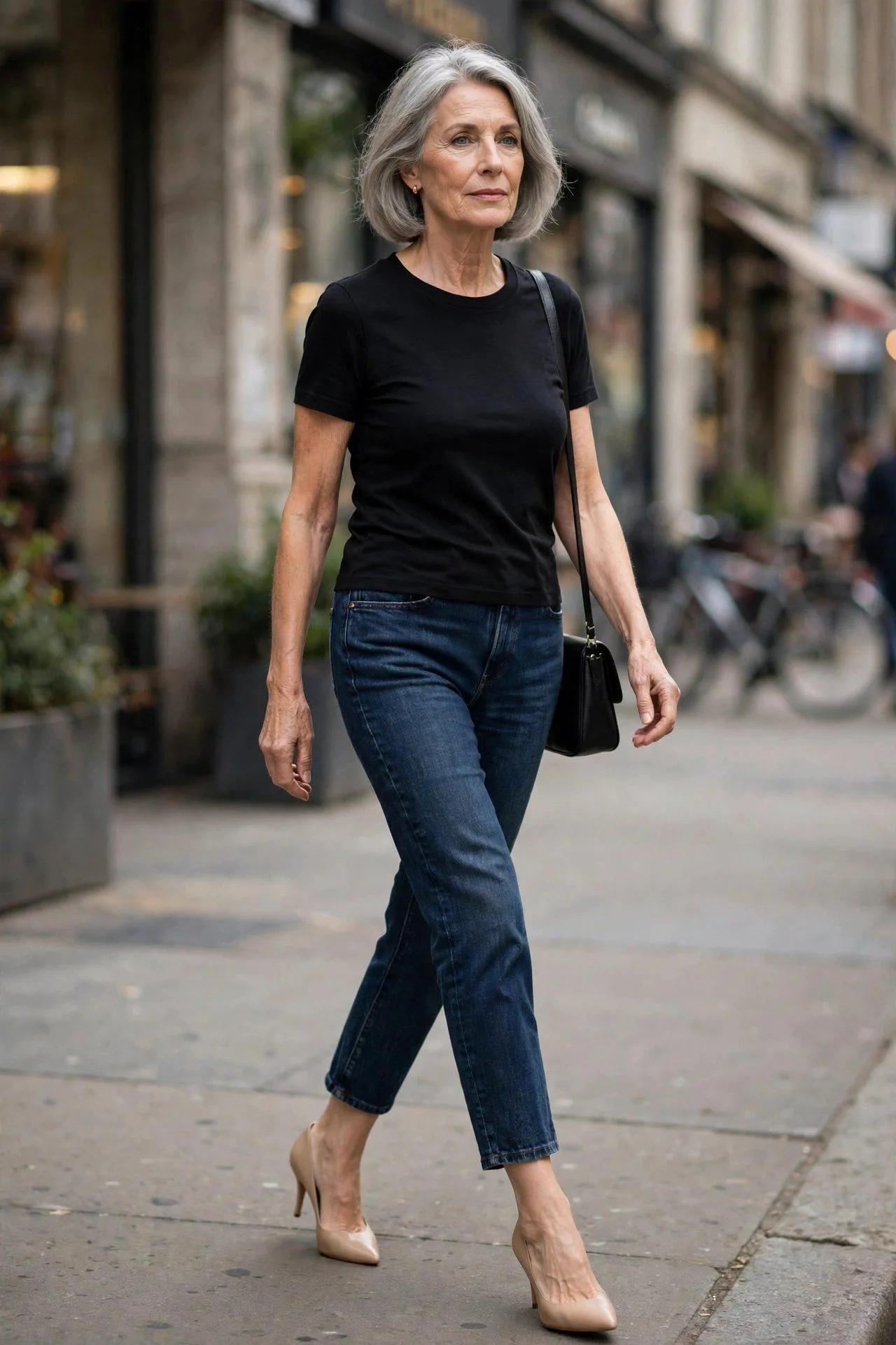 Grey-bobbed woman striding through the city in dark cropped jeans, a black top, and classic nude pointed-toe pumps.